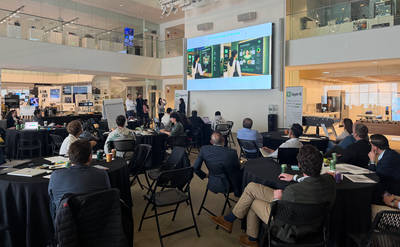Participants seated at round tables in the MIT Media Lab watch a presentation on a large wall-mounted screen during a TD Bank hackathon event.