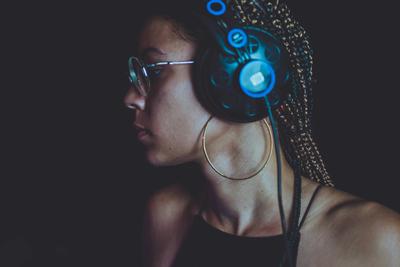 A person with braided hair, round glasses, and hoop earrings listens intently through illuminated headphones against a dark background.